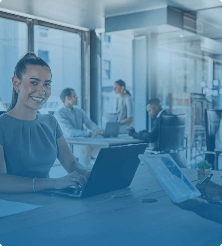 A woman in a professional setting smiling at her laptop with colleagues working in the background.