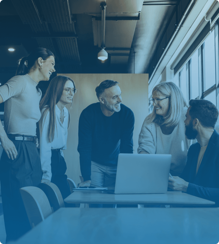 A group of professionals in a collaborative meeting, standing around a laptop and smiling.