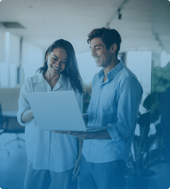 Two colleagues laughing and discussing work while looking at a laptop in a modern office setting.