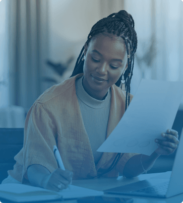 A woman working at a desk, holding a report while writing with a pen, indicating data analysis, reporting, or content review