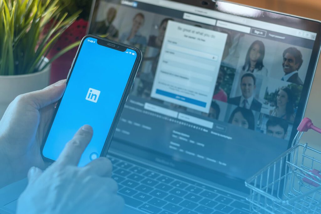 Person using the LinkedIn mobile app with the LinkedIn login screen visible on a laptop in the background