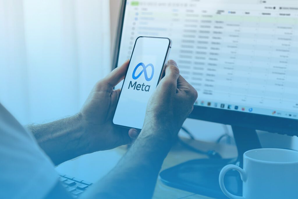 Person holding a smartphone displaying the Meta logo in front of a desktop screen with spreadsheets