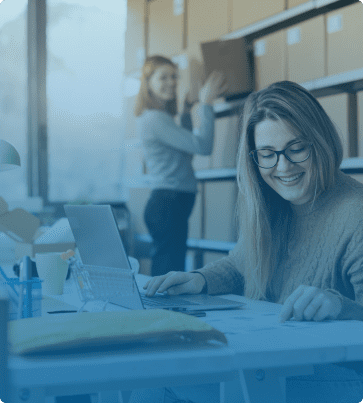 Ecommerce SEO Image: A woman sitting at a desk in a warehouse setting, smiling while working on documents and a laptop, with shelves of boxes in the background.
