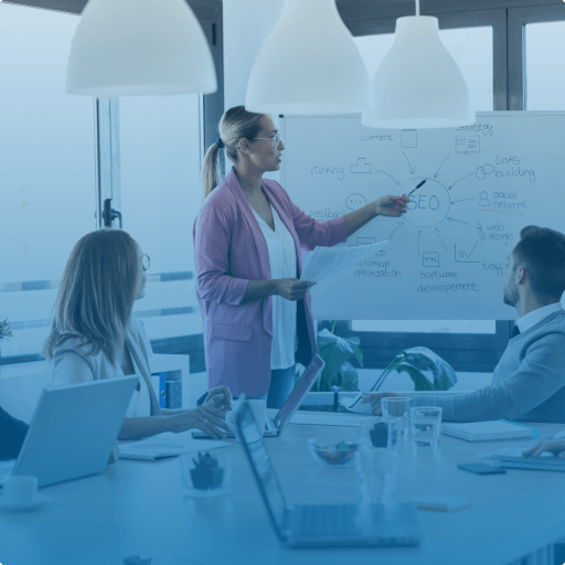 A team in a meeting room discussing an SEO strategy, with a woman presenting a diagram on a whiteboard